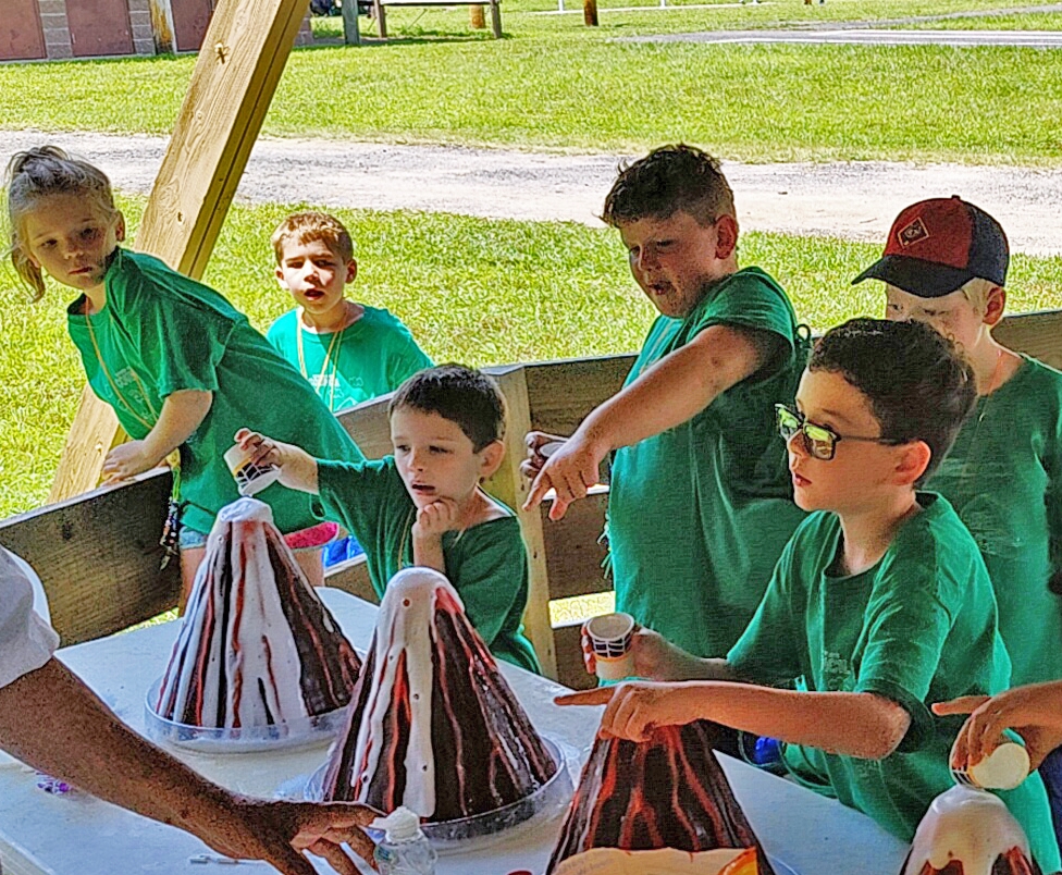 Four elementary aged kids create baking soda volcanoes at an outdoor day camp while other kids look on in excitement