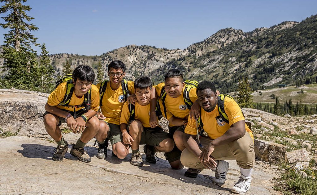 5 teen boys of diverse cultural heritage wear matching yellow t-shirts and pose, crouching, on a scenic mountain top hike on a clear, sunny day
