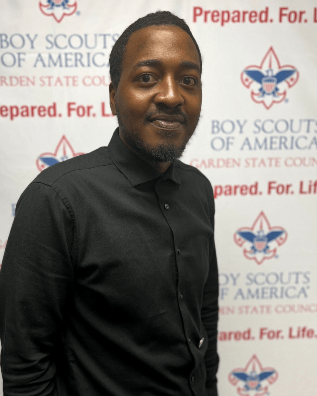 Maurice Bullock is a young man with dark brown skin wearing a black shirt. He stands with a warm, confident expression in front of a white background with repeating blue and red logos for the Boy Scouts of America Garden State Council