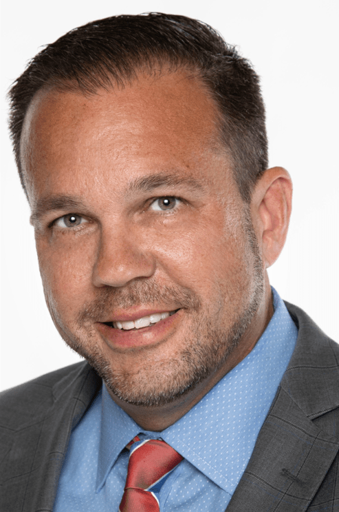 Tim Guim is a man with light skin and brown hair and face stubble who is smiling and wearing light blue dress shirt, red tie, and grey jacket.